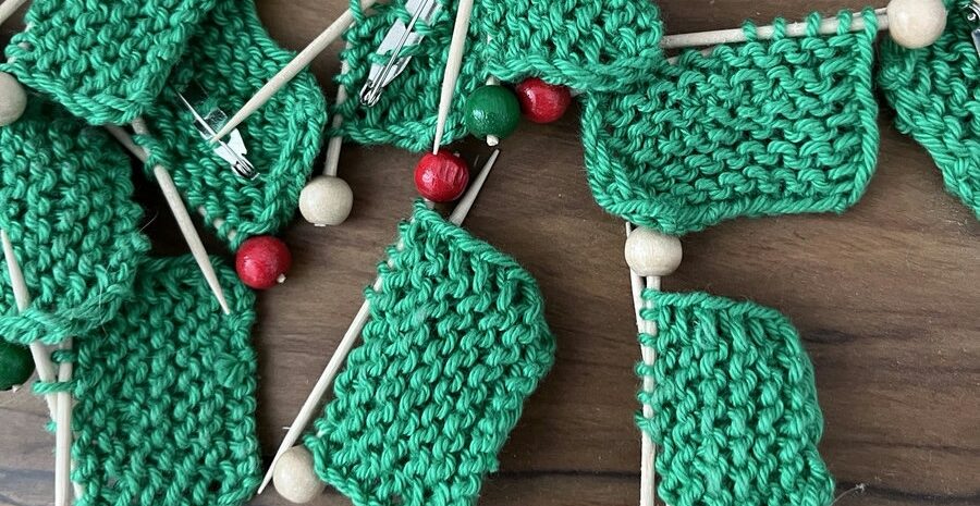 Handgestrickte grüne Anstecker in Form von Mini-Strickzeug: kleine Strickstücke auf Holznadeln mit roten, grünen und hellen Holzperlen.