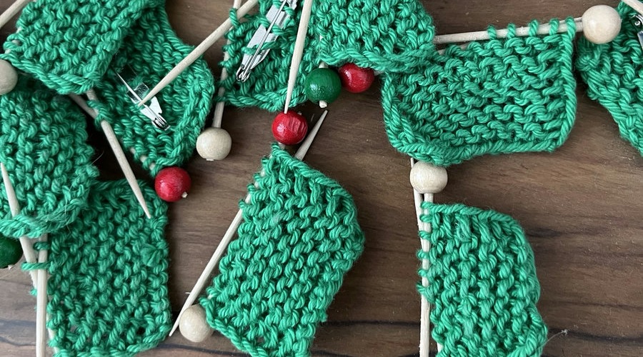 Handgestrickte grüne Anstecker in Form von Mini-Strickzeug: kleine Strickstücke auf Holznadeln mit roten, grünen und hellen Holzperlen.